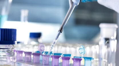 An analytical scientist's hand holds a pipette to prepare a sample for elemental analysis services, pipetting liquid into purple and blue sample vials.